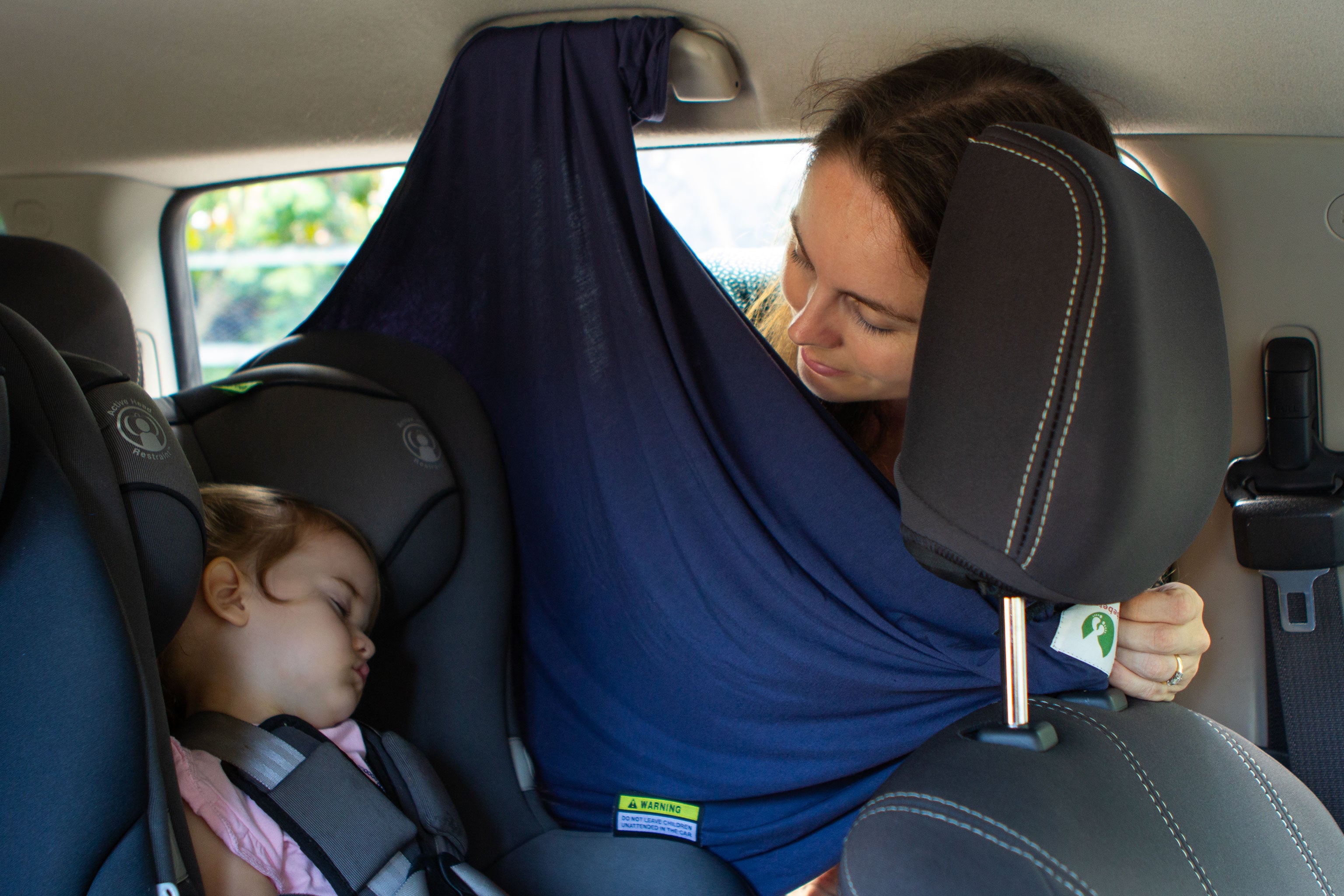 A mother looks at her sleeping baby in her car seat, protected from the sun with a Bebe Trek Multifunctional magnetic sun cover hanging from the grab handle